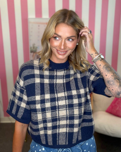 A woman wearing a navy and cream plaid short-sleeve knit sweater, standing in front of a pink and white striped wall.