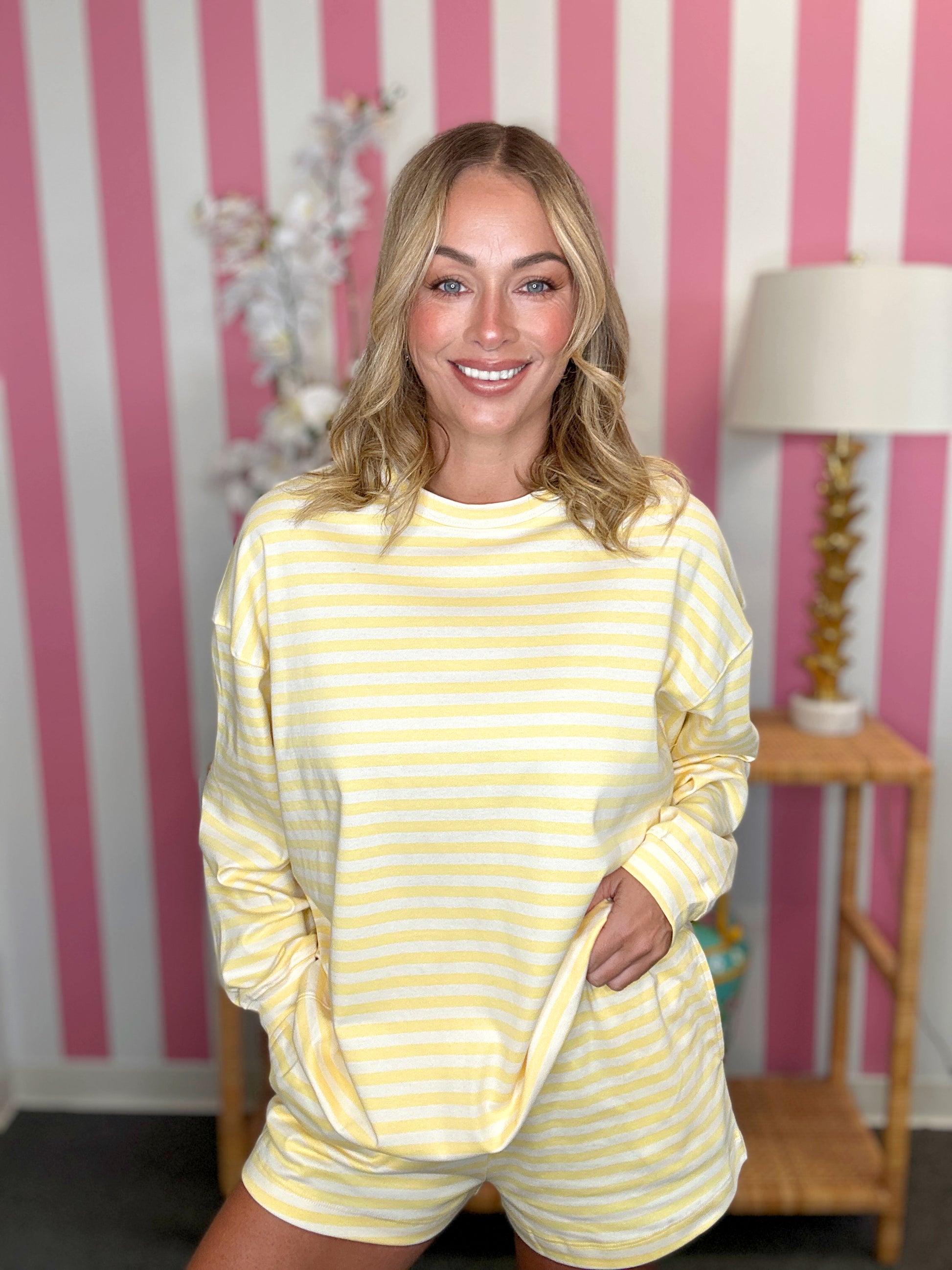 A smiling blonde woman wearing a yellow and white striped long-sleeve top and matching shorts, standing in front of a pink and white striped wall with a lamp on a side table in the background.