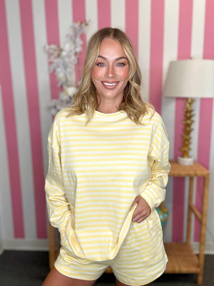 A smiling blonde woman wearing a yellow and white striped long-sleeve top and matching shorts, standing in front of a pink and white striped wall with a lamp on a side table in the background.