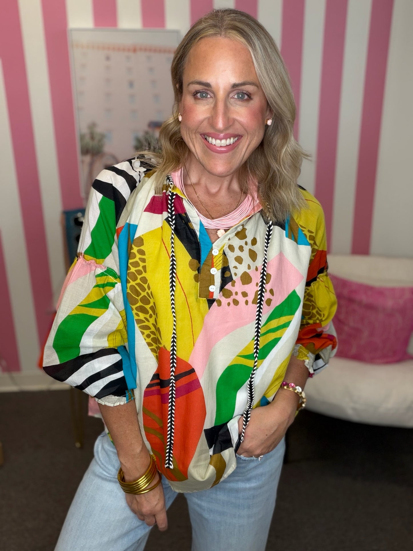A smiling blonde woman wearing a vibrant, multicolored abstract-patterned blouse and light blue jeans, standing in front of a pink and white striped wall.