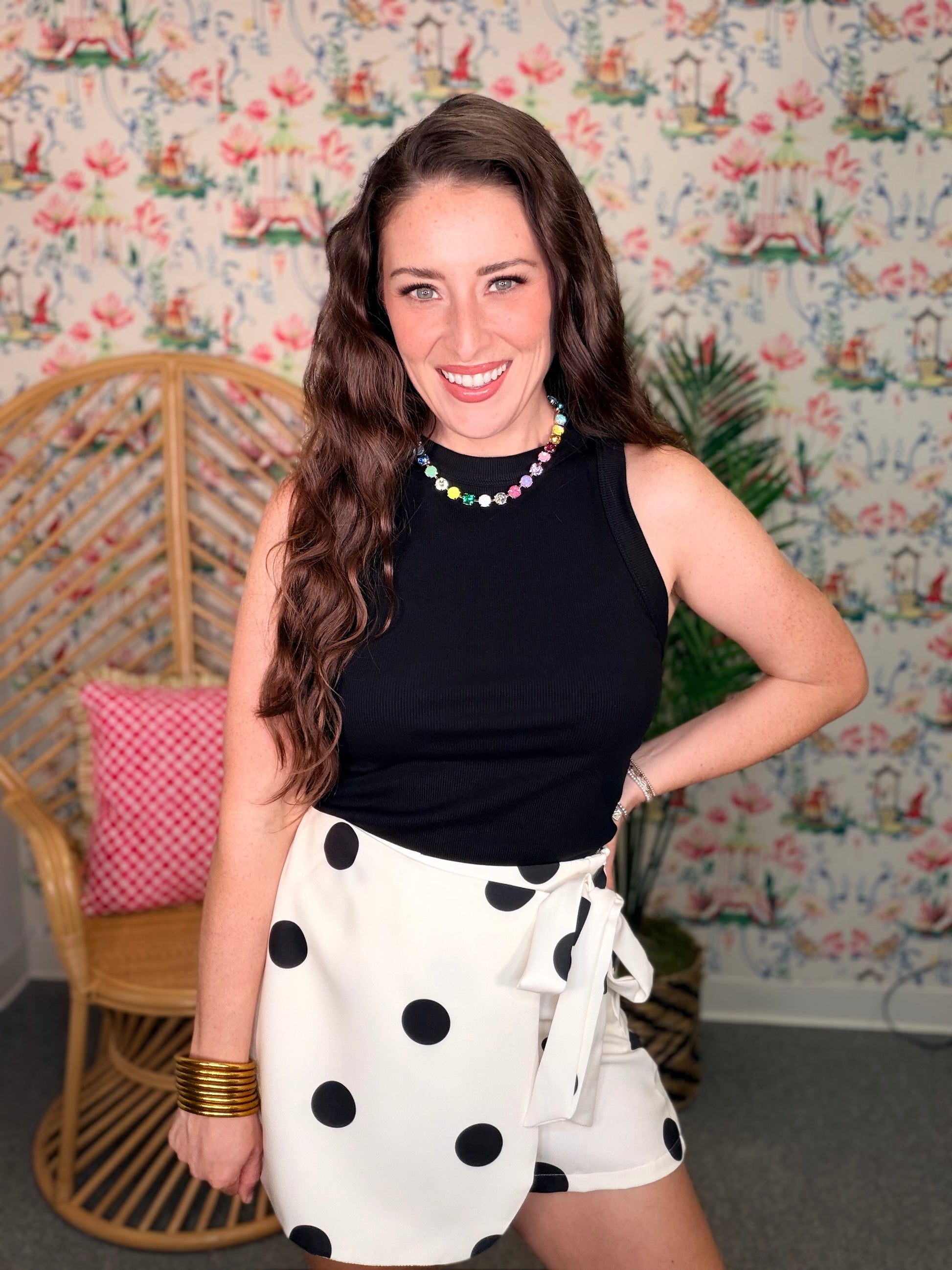 Woman wearing a black sleeveless top and white polka dot skirt in a room with floral wallpaper.