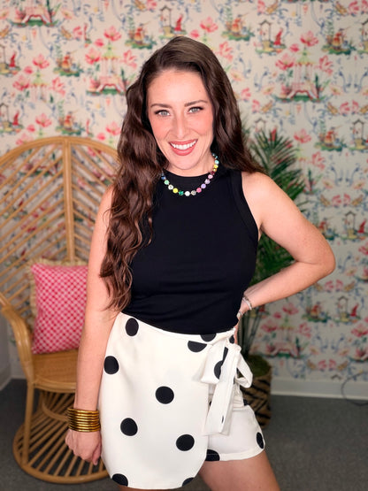 Woman wearing a black sleeveless top and white polka dot skirt in a room with floral wallpaper.