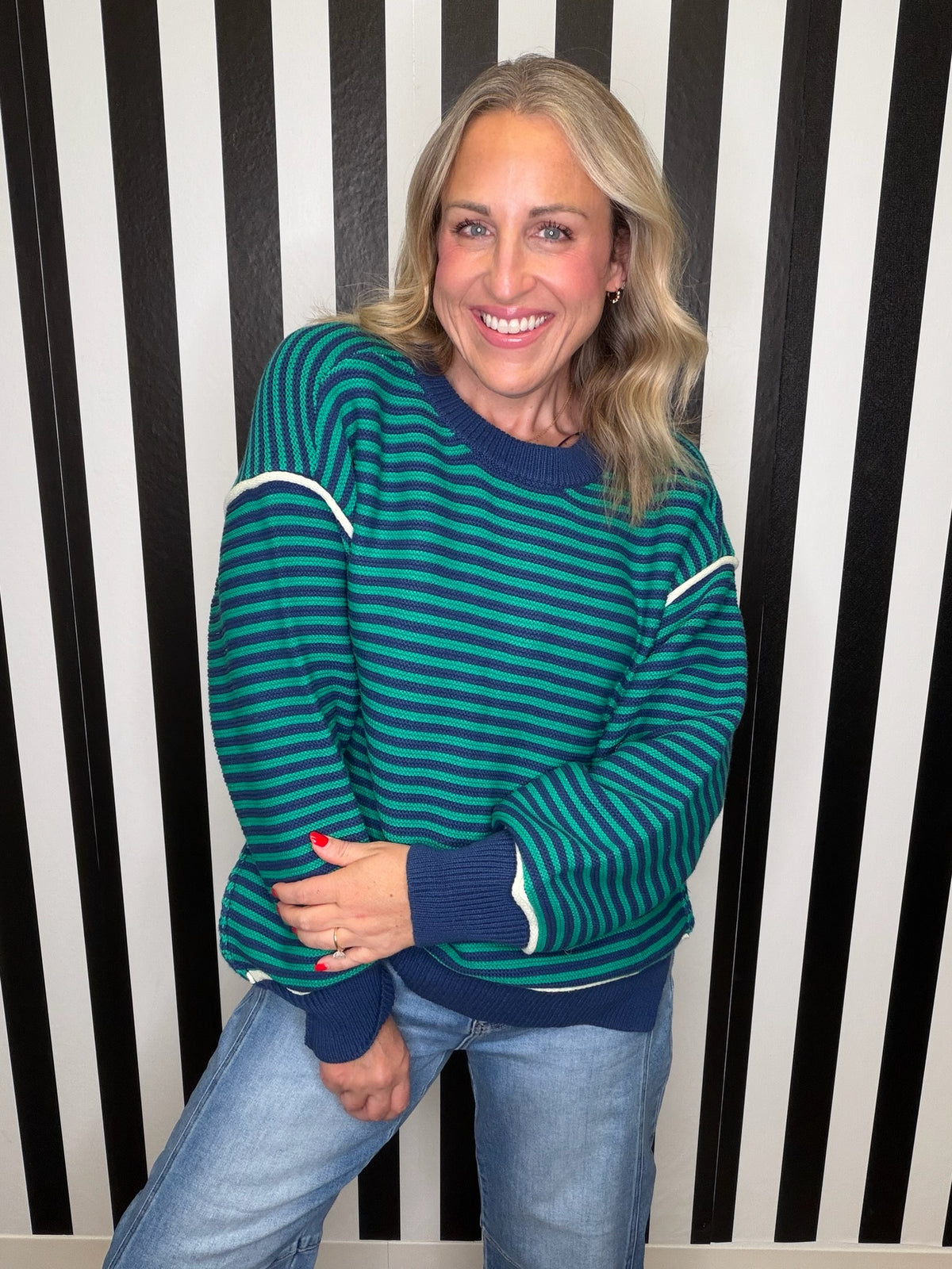 A smiling woman wearing a teal and navy striped sweater with white contrast stripes on the shoulders and cuffs, paired with light blue jeans, standing in front of a black and white vertically striped wall.