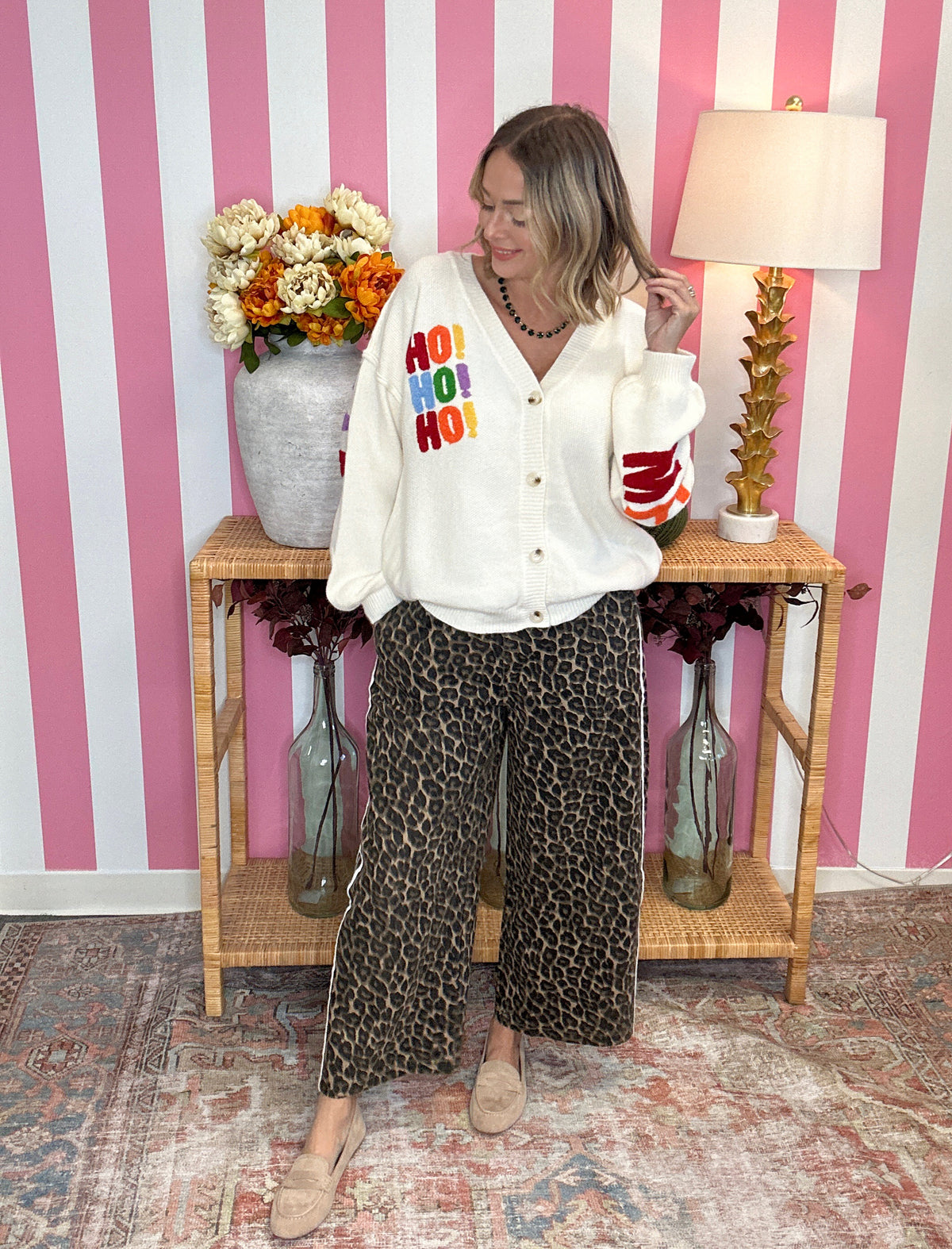 A woman stands in a room with pink and white striped walls, wearing a white button-up cardigan with colorful 'HO HO HO' text on the chest and leopard-print wide-leg pants.