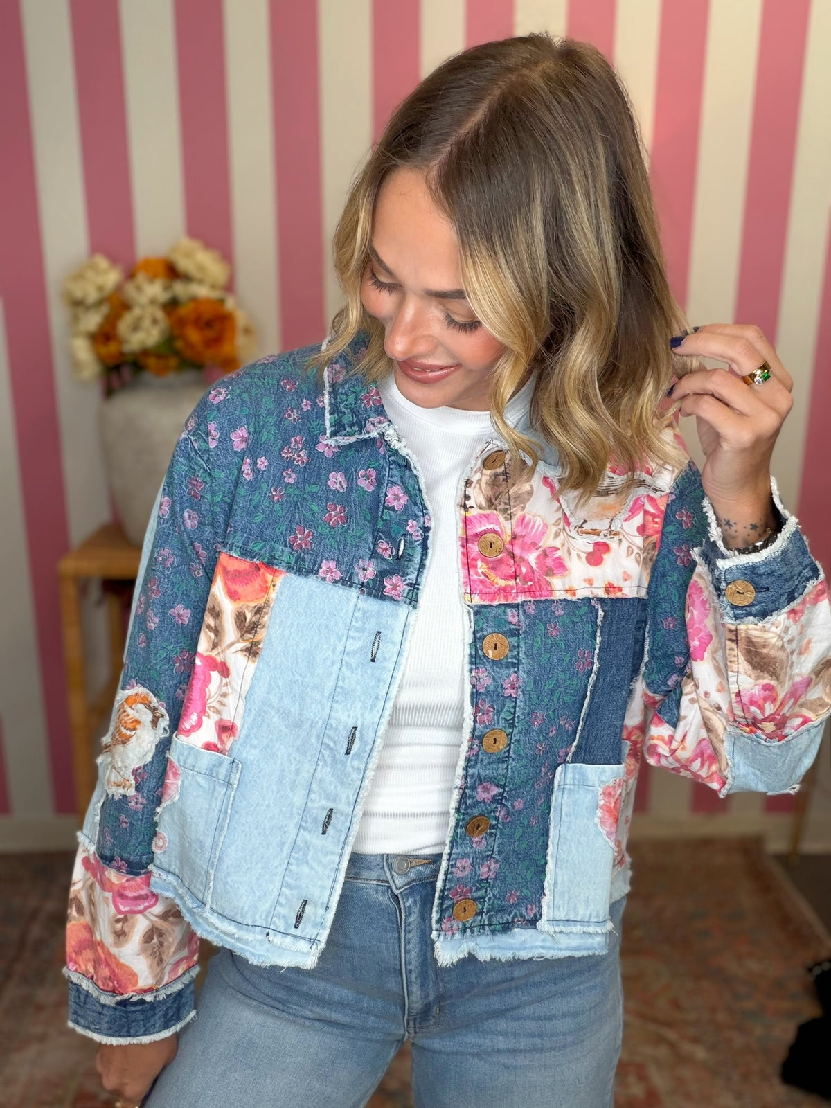A woman wearing a patchwork denim jacket with floral prints, a white t-shirt, and light-wash jeans, standing in front of a pink and white striped wall.
