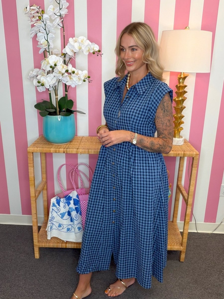 A woman wearing a blue and white gingham sleeveless midi dress stands in front of a pink and white striped wall, next to a wooden side table with a turquoise orchid pot and a lamp.