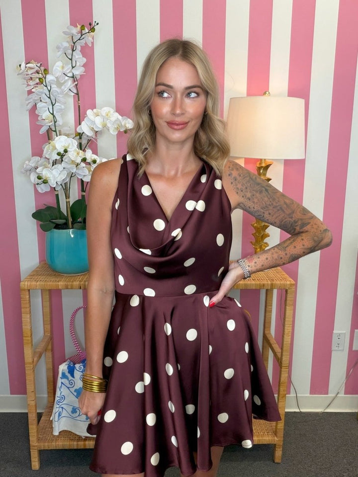 A woman modeling a sleeveless cowl-neck fit-and-flare dress in dark brown with large white polka dots, standing in front of a pink and white striped wall.
