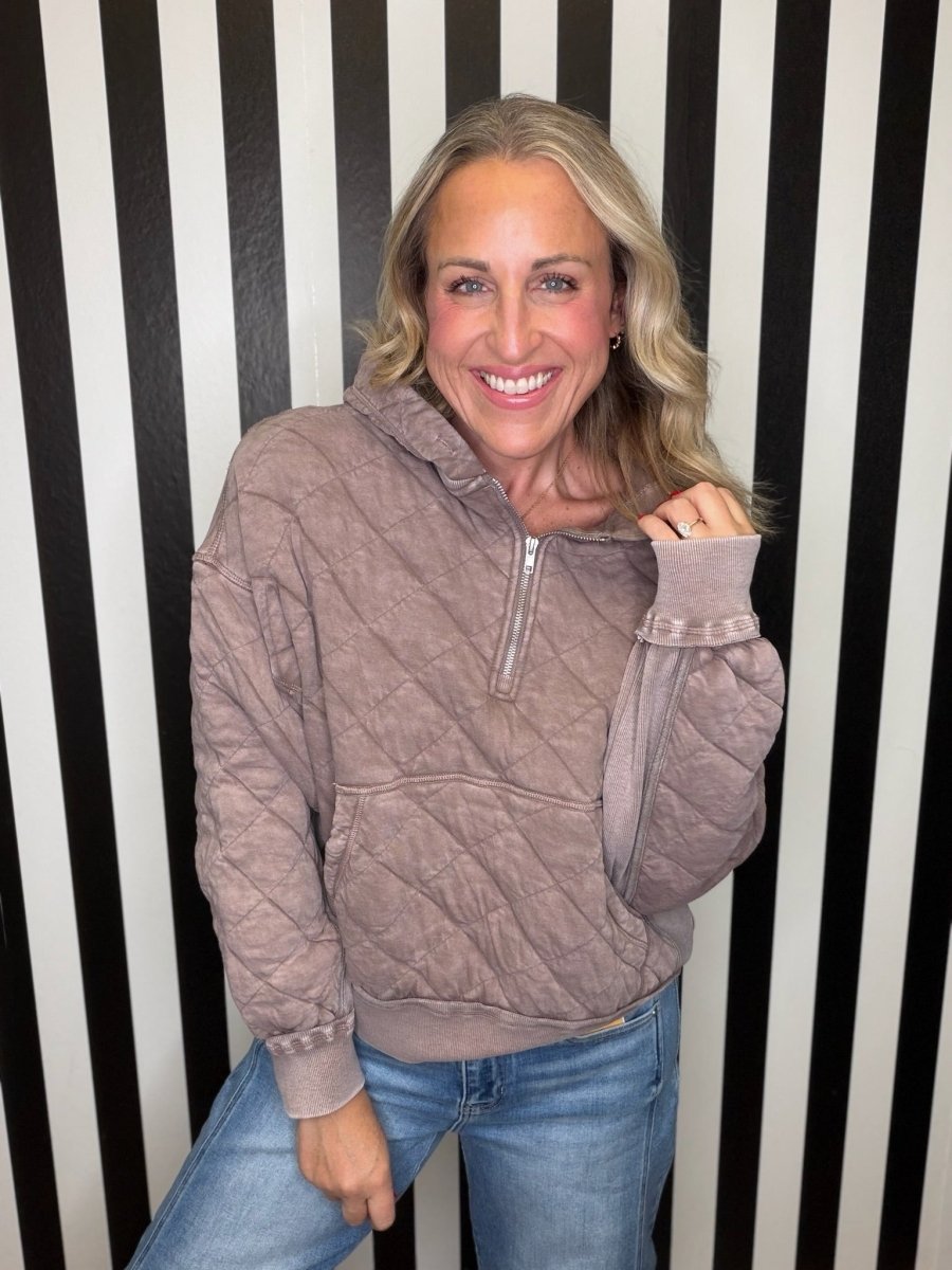 A smiling blonde woman wearing a taupe quilted half-zip hoodie and blue jeans, posing in front of a black and white vertically striped wall.