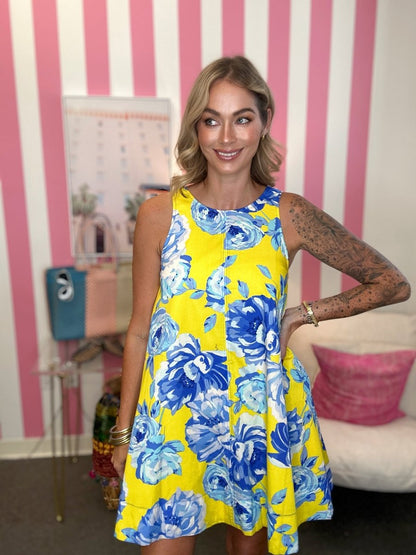 A woman wearing a sleeveless yellow A-line dress with a blue and light blue floral print, standing in front of a pink and white striped wall.