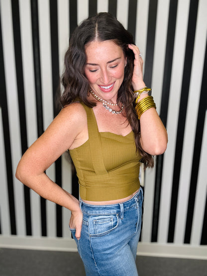 A woman posing in a ribbed olive green crop top and blue jeans, accessorized with gold bangles and a silver necklace, set against a black and white striped wall.