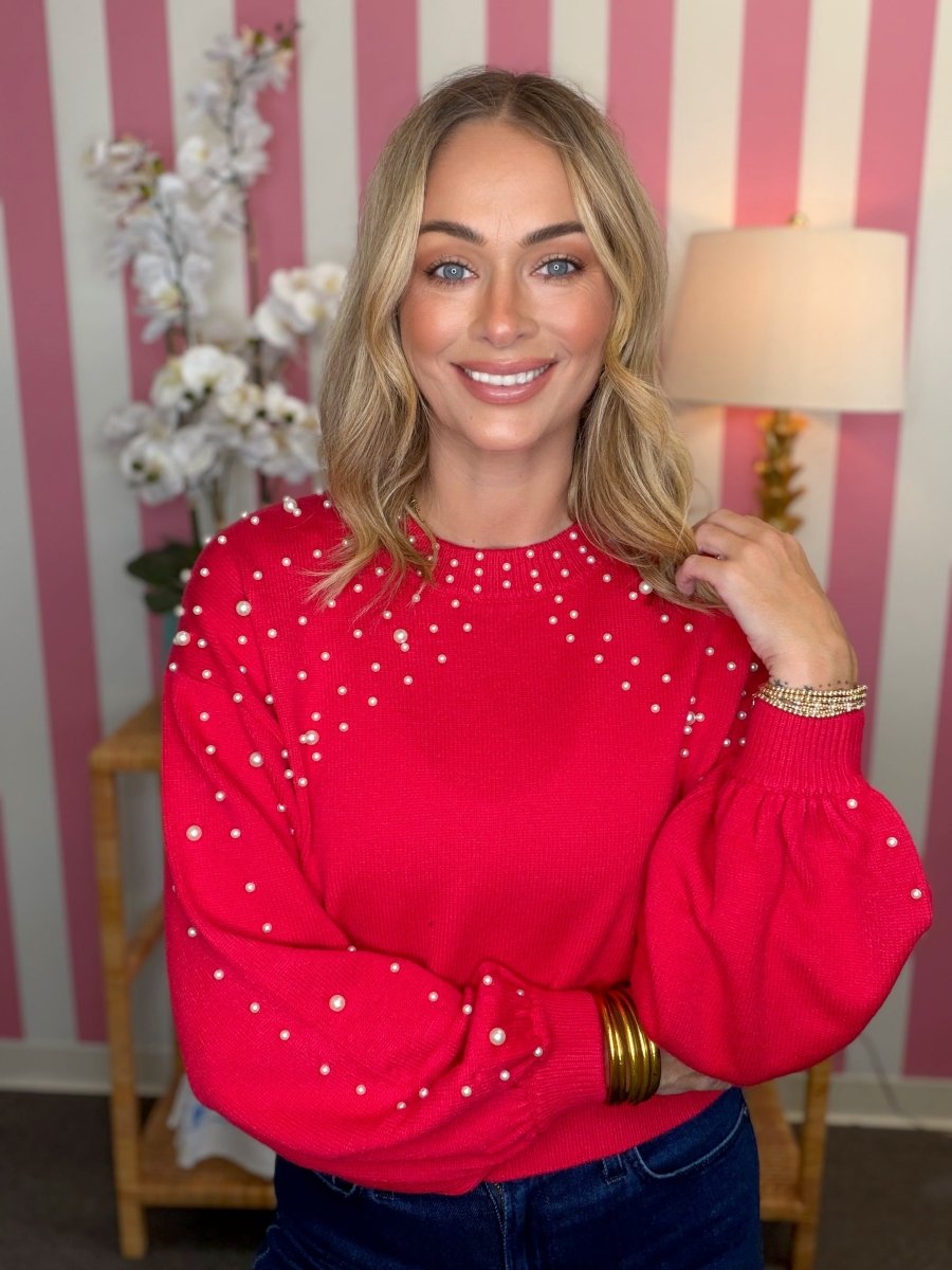 A smiling blonde woman wearing a bright red knit sweater with white pearl-like embellishments, posed in front of a pink and white striped wall with a lamp and flowers in the background.