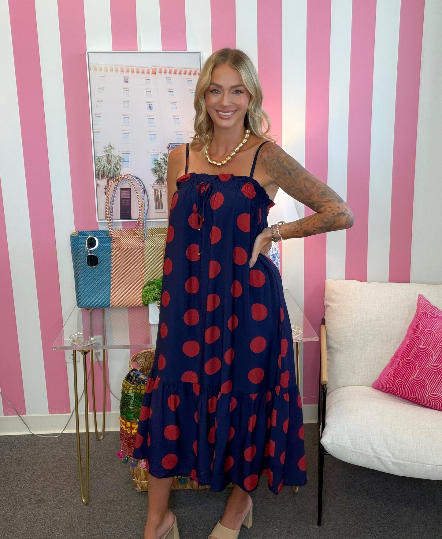 A woman modeling a navy blue midi dress with red polka dots, standing in front of a pink and white striped wall. She is wearing a gold beaded necklace and beige heels, with a glass table and a white sofa visible in the background.