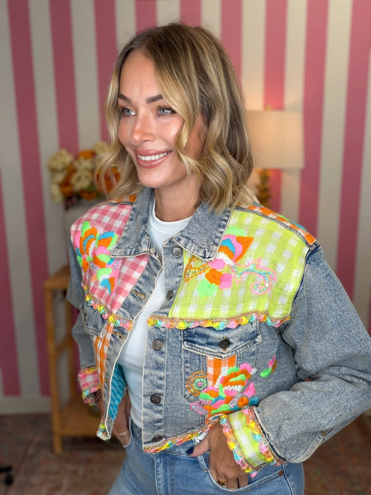 A woman wearing a denim jacket with vibrant, multicolored patchwork fabric panels on the shoulders and front pocket, styled with a white t-shirt and jeans against a pink and white striped wall.