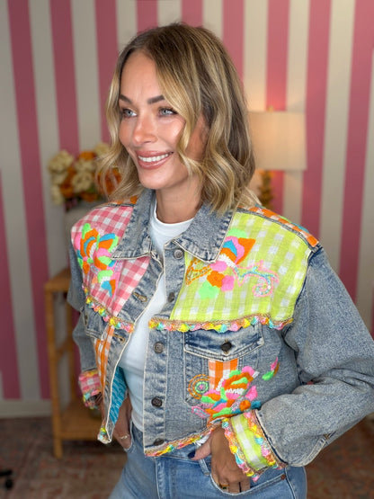 A woman wearing a denim jacket with vibrant, multicolored patchwork fabric panels on the shoulders and front pocket, styled with a white t-shirt and jeans against a pink and white striped wall.