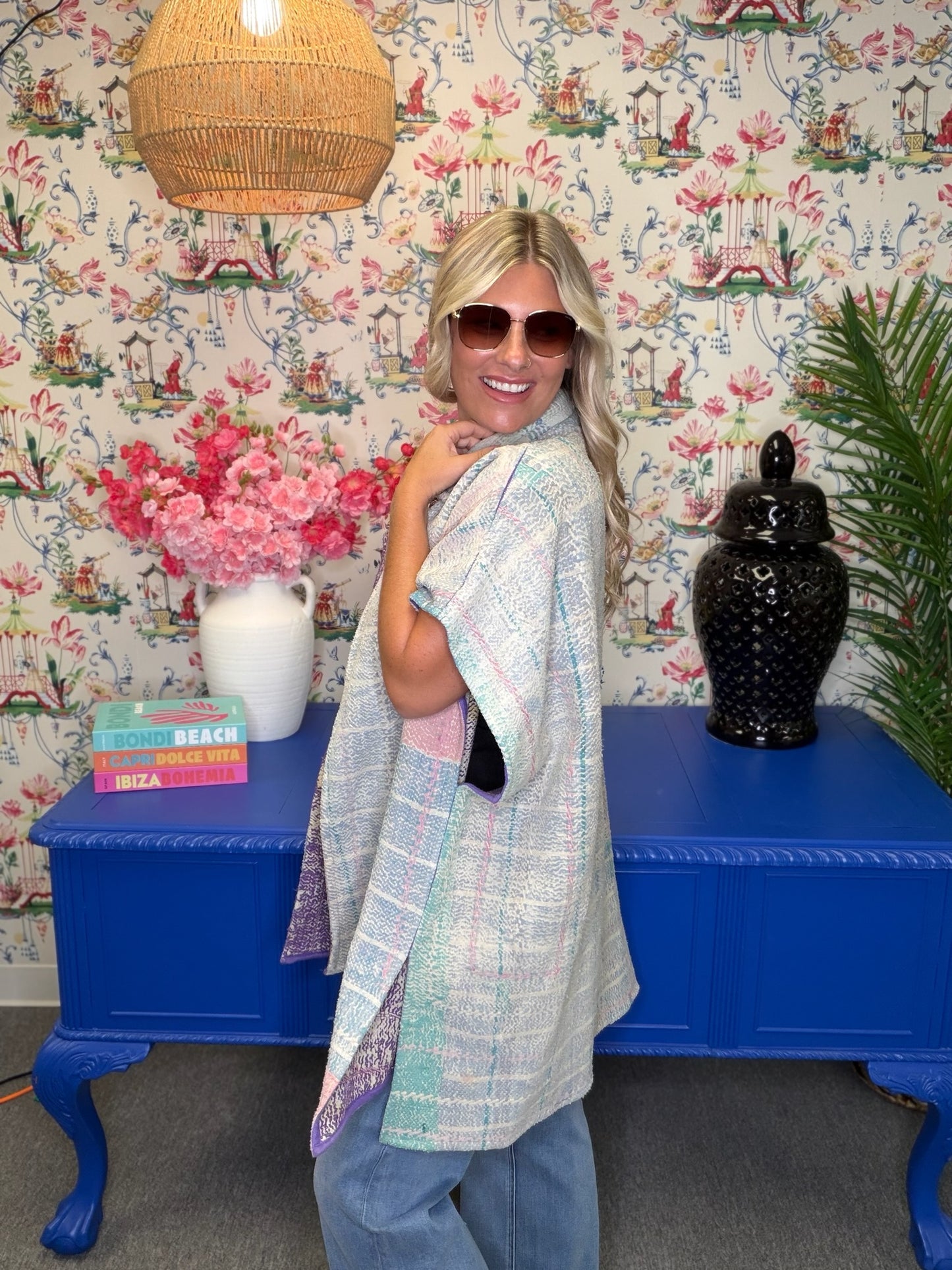 A woman wearing a pastel multicolor plaid tunic top and light blue jeans stands in front of a blue console table decorated with pink flowers, a black ceramic jar, and tropical wallpaper.