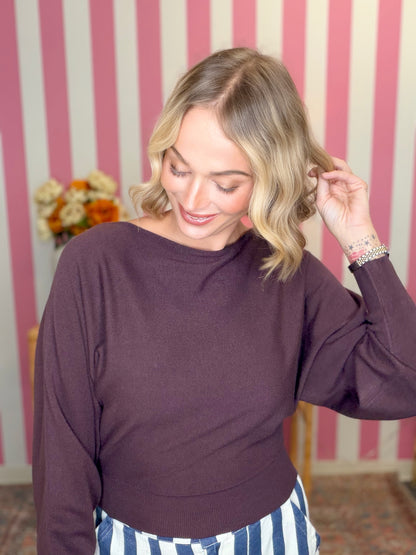 A woman with blonde wavy hair wearing a dark plum purple long-sleeve knit top and blue-and-white striped pants, posing in front of a pink and white striped wall.