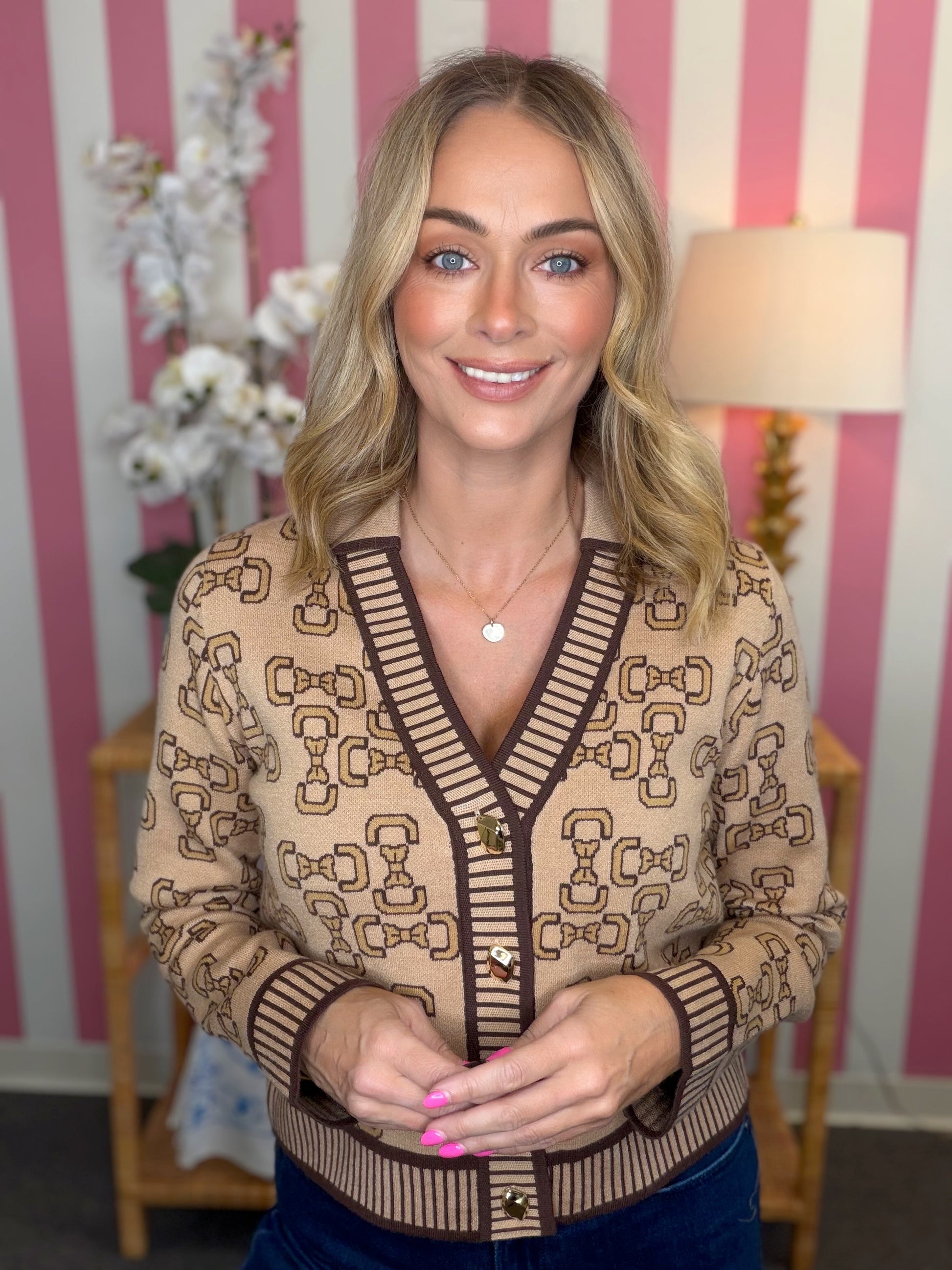 A smiling blonde woman wearing a beige and brown patterned knit cardigan sweater with dark brown trim and gold buttons, standing in front of a pink and white striped wall.