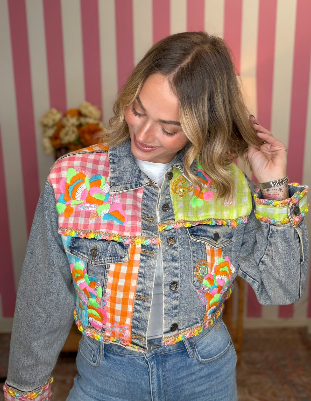 A woman wearing a heavily customized denim jacket with colorful candy-themed patchwork panels, posing in front of a pink and white striped wall.