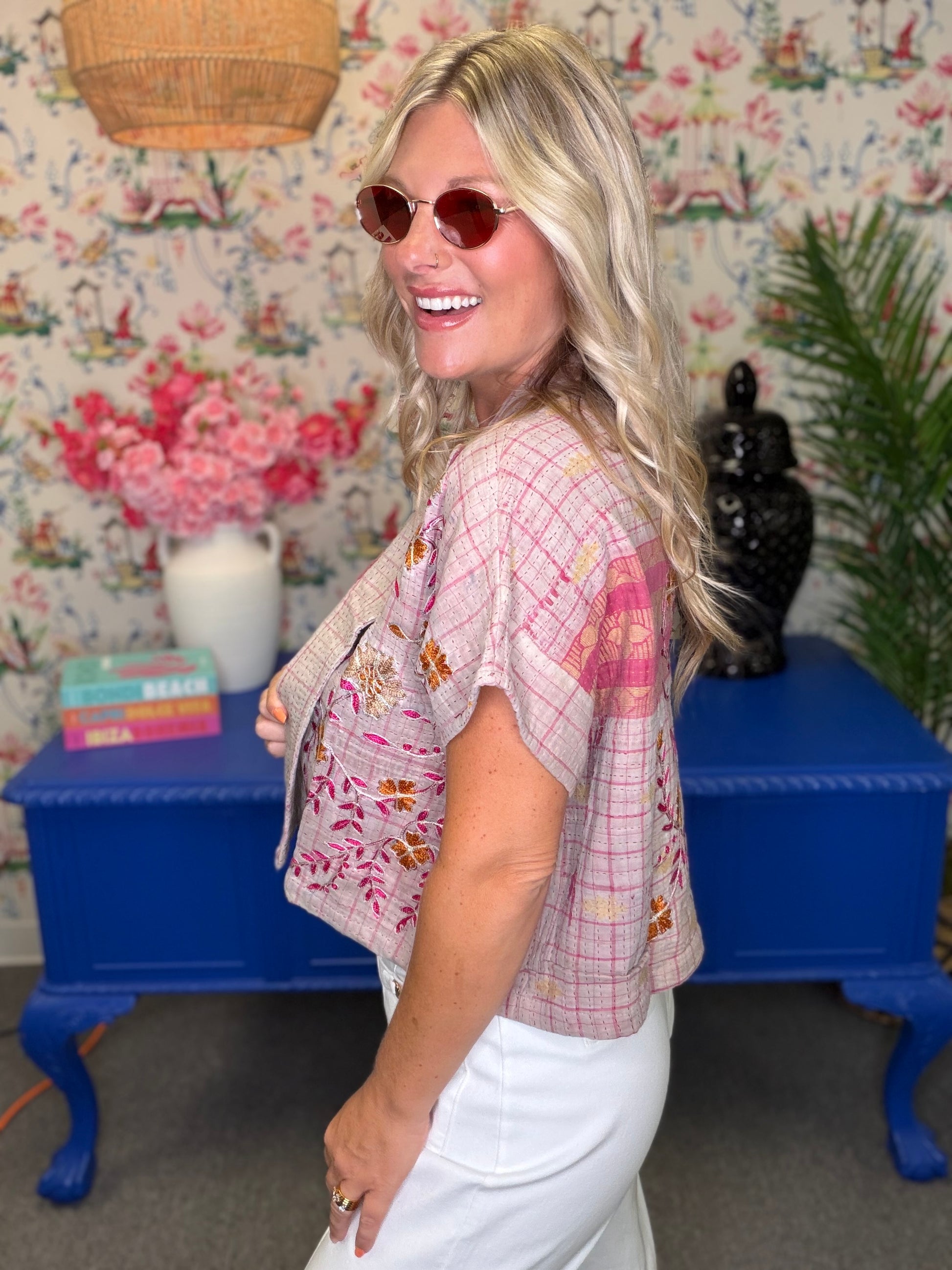 A woman with blonde hair and round sunglasses is shown from the side/back, wearing a light pink plaid short-sleeve crop top with a floral print overlay and white pants, standing in a decorated room with a blue side table and floral wallpaper.