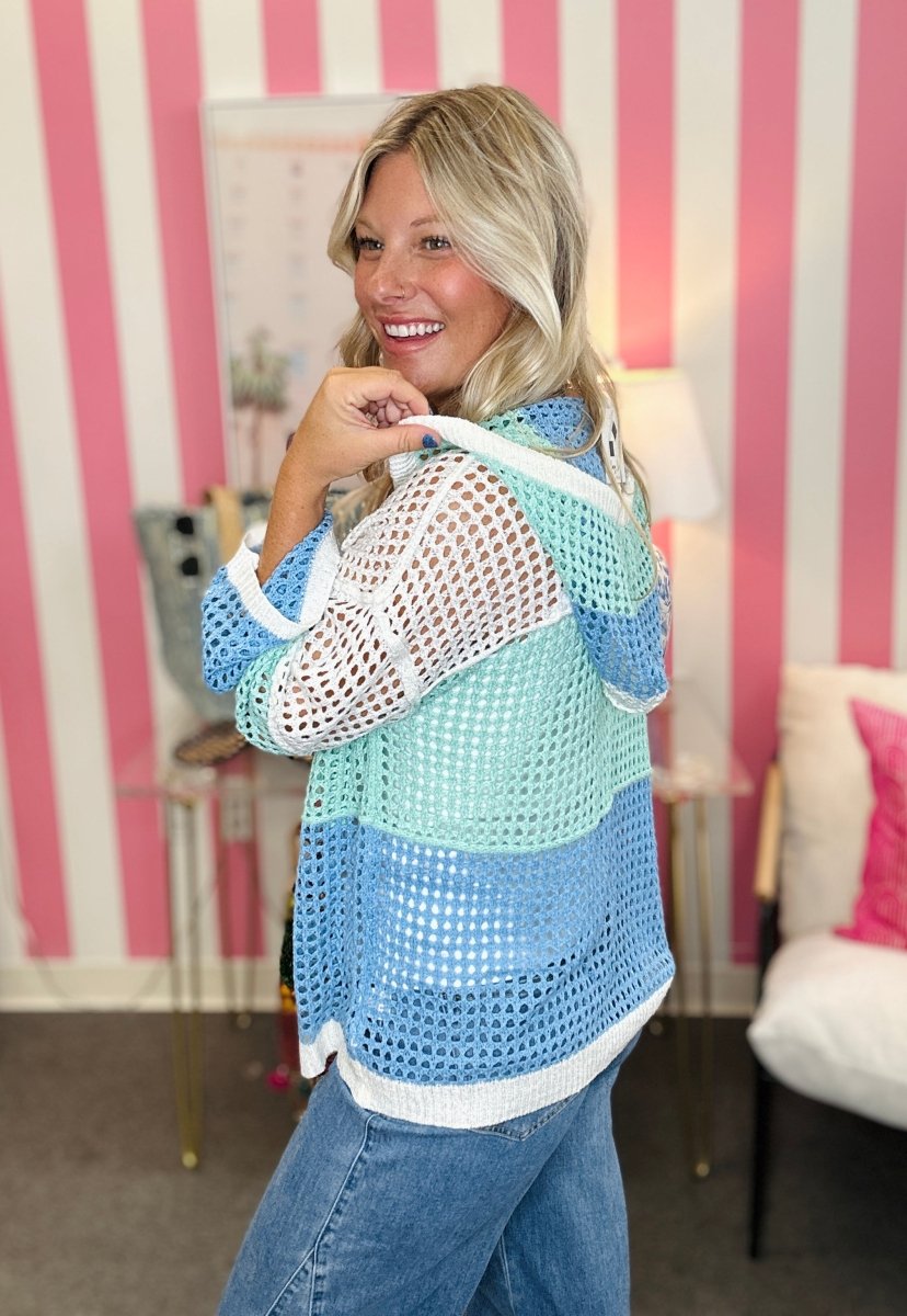 A woman wearing a colorful striped crochet cardigan with blue, mint green, and white stripes, paired with blue jeans, standing in a room with pink and white striped wallpaper.