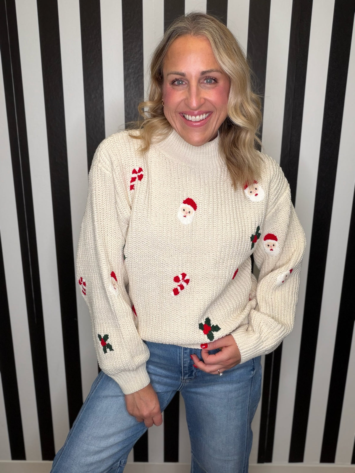 A smiling blonde woman wearing a cream-colored knit sweater with red and green Christmas motifs, paired with blue jeans, standing in front of a black and white striped wall.