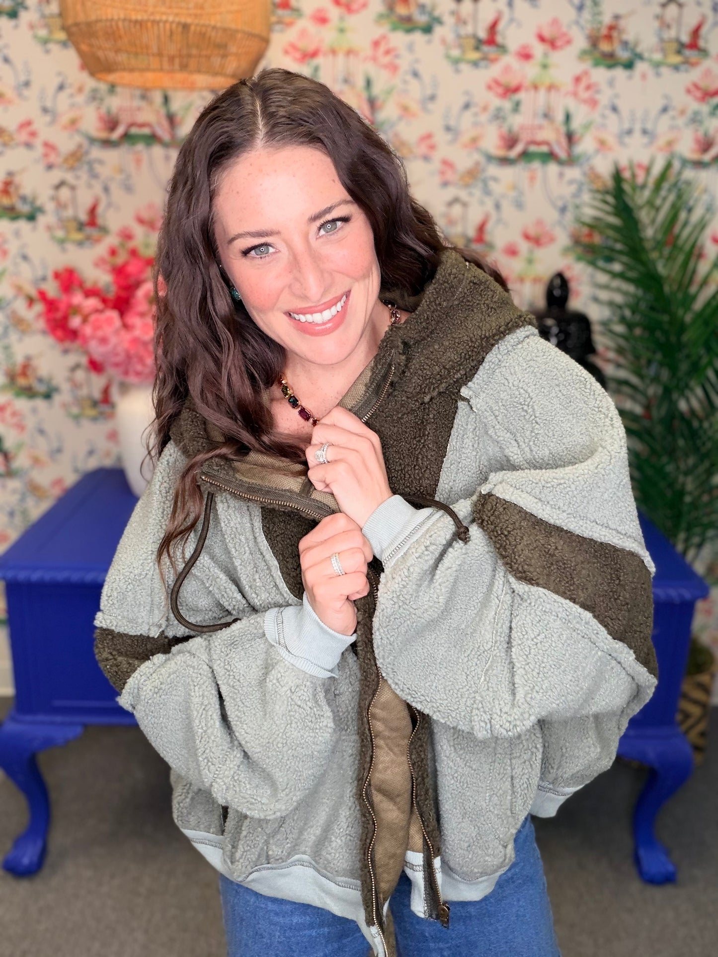 A woman smiling while wearing a chunky fleece hooded jacket with sage green and dark brown color-blocked panels, standing in front of a floral wallpaper background with a blue table.