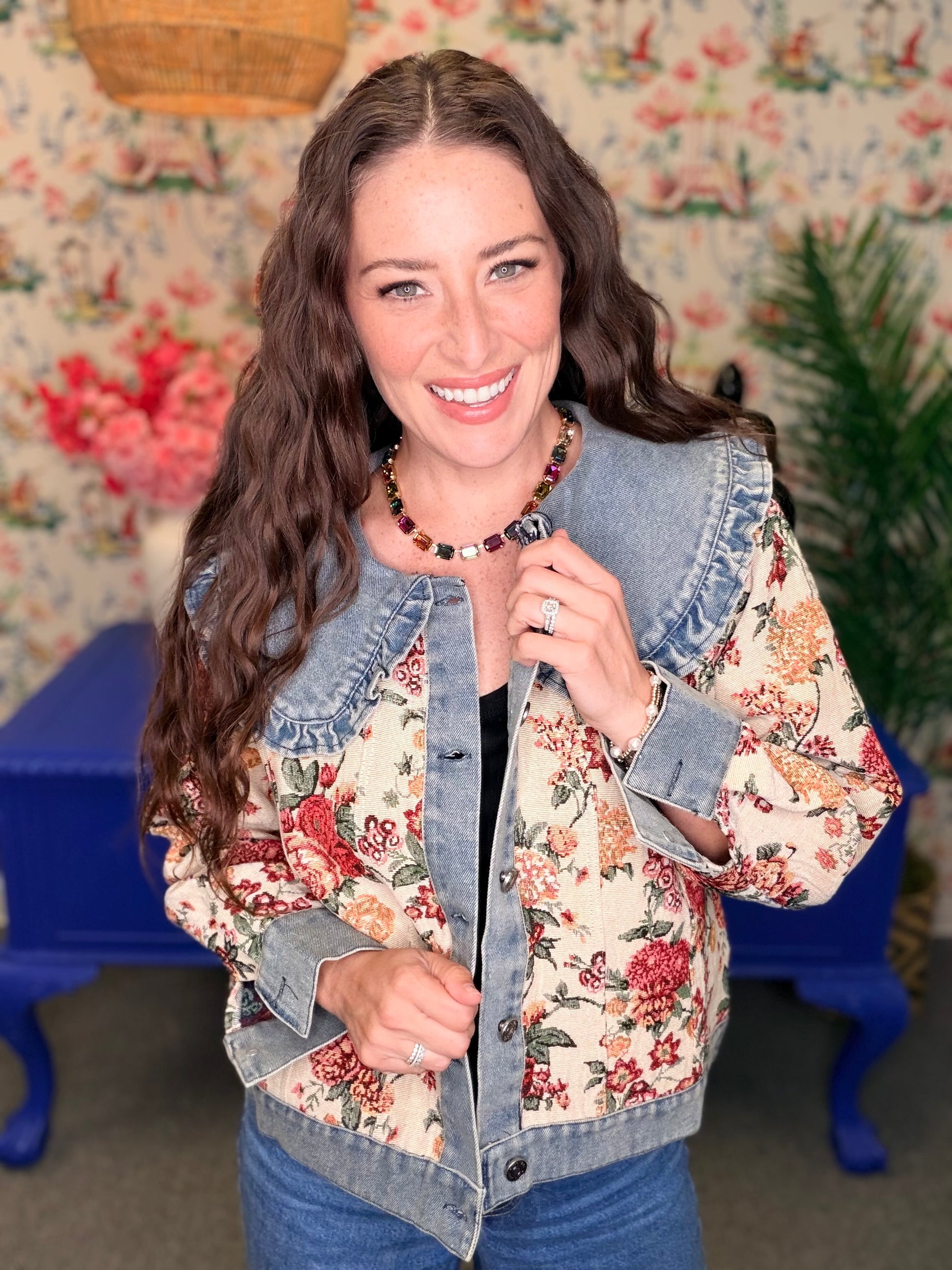 A woman smiling and wearing a denim jacket with a red and orange floral print on the front panels, set against a vintage floral wallpaper background with a blue table.
