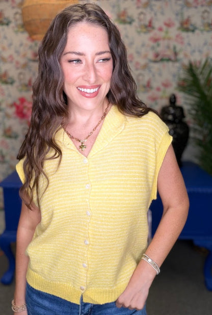 A woman with long wavy brown hair smiling, wearing a yellow knit short-sleeve button-up top and blue jeans, standing in front of a floral wallpaper background.