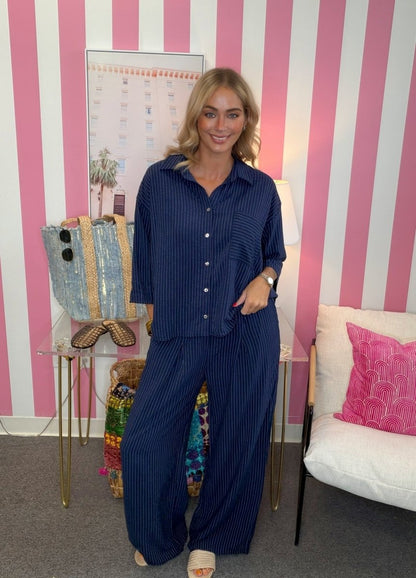 A woman modeling a matching navy blue pinstriped button-up shirt and wide-leg trousers set, standing in a room with pink and white vertical stripes.
