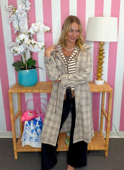 A woman models a long beige and brown plaid coat over a striped shirt and dark pants, standing in front of a pink and white striped wall next to a wooden console table with a lamp and flowers.