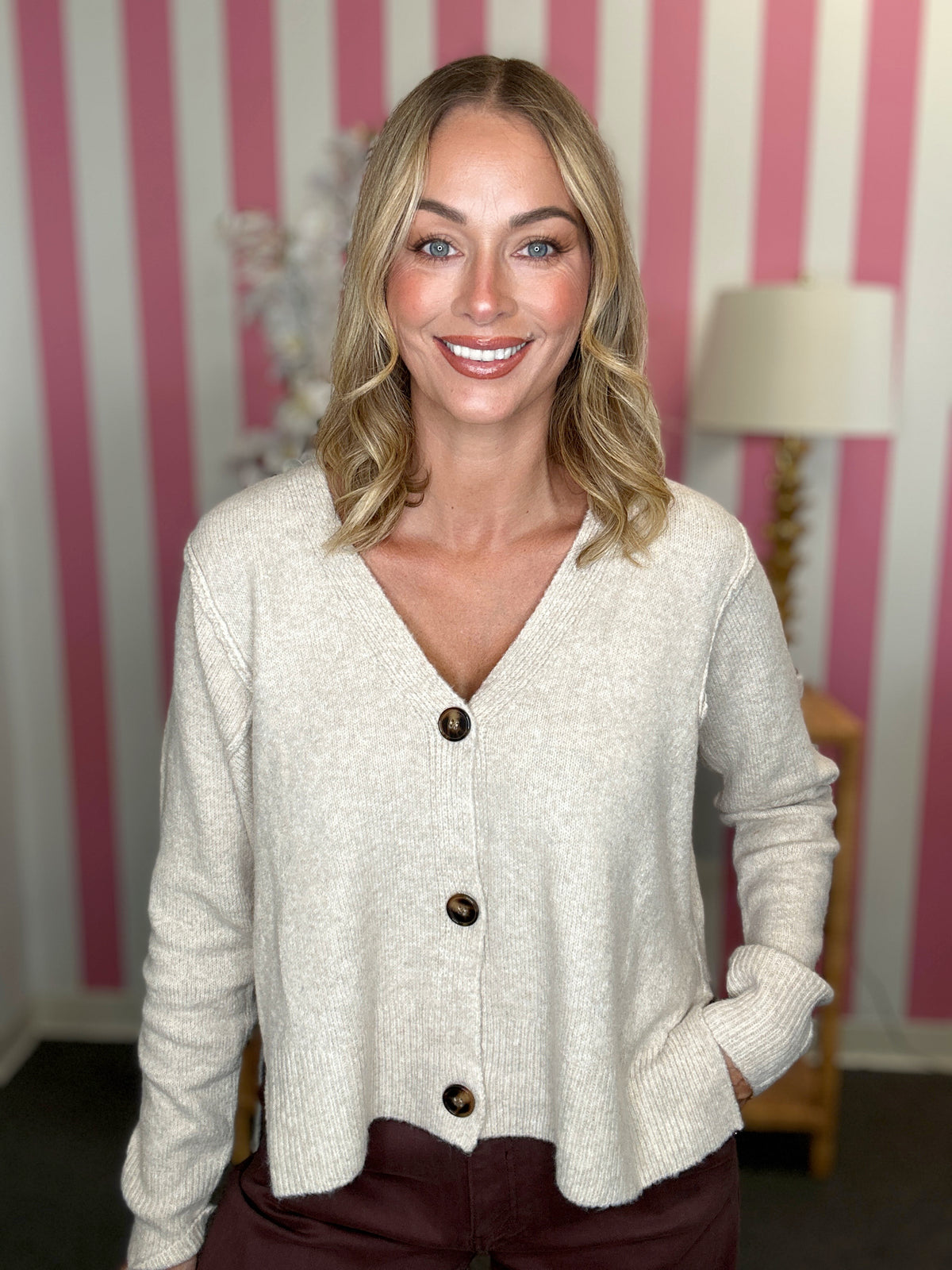 A smiling blonde woman wearing a light oatmeal knit cardigan with dark buttons and dark brown pants, standing in front of a pink and white striped wall.