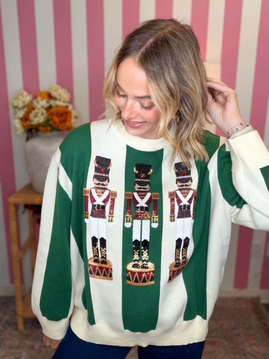 A woman wearing a green and cream knit sweater with three large nutcracker graphics on the front, posed in front of a pink and white striped wall.