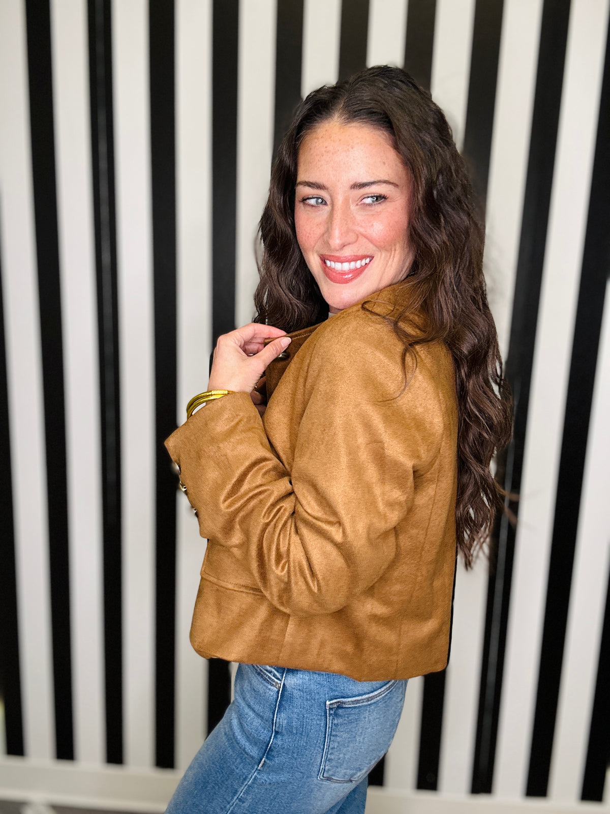 A woman with long wavy brown hair is shown from the back wearing a cropped tan faux-leather jacket and blue jeans, posing in front of a black and white vertically striped background.