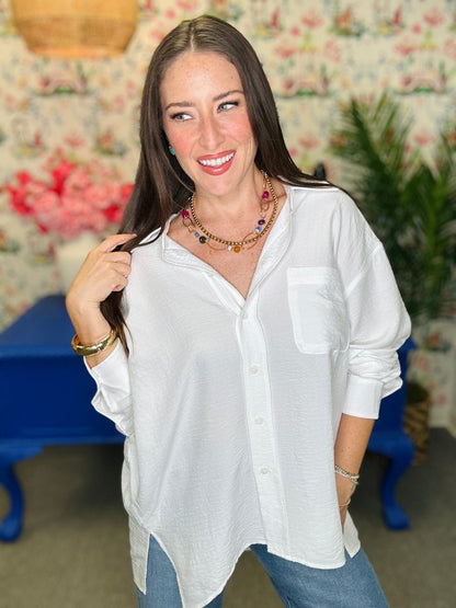 A woman wearing a white long-sleeve button-down shirt with a chest pocket, paired with blue jeans, standing in a room with floral wallpaper and a blue bench in the background.