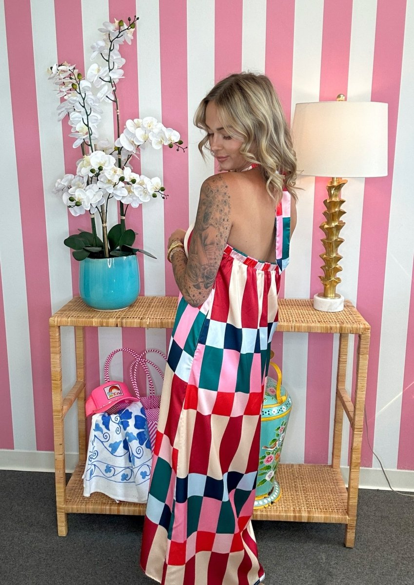 A woman wearing a colorful patchwork checkered maxi dress stands in front of a pink and white striped wall, next to a wicker console table with a lamp and flowers.