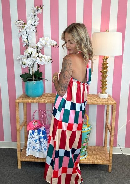 A woman wearing a colorful patchwork checkered maxi dress stands in front of a pink and white striped wall, next to a wicker console table with a lamp and flowers.