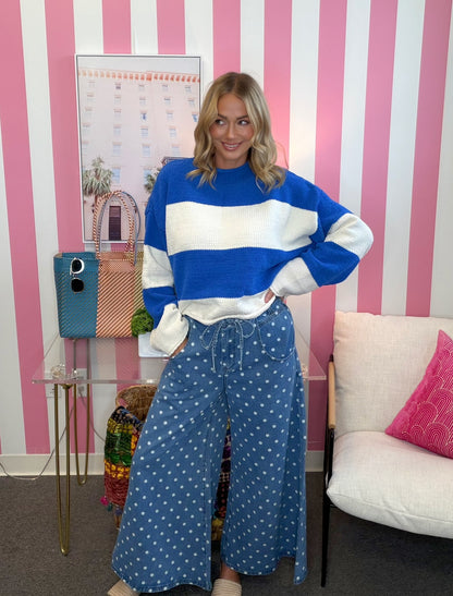 A woman stands in a room with pink and white striped walls, wearing a blue and white striped crewneck sweater and blue wide-leg pants with white polka dots. She is posing next to a white chair with a pink pillow and a glass side table holding a blue and pink handbag.