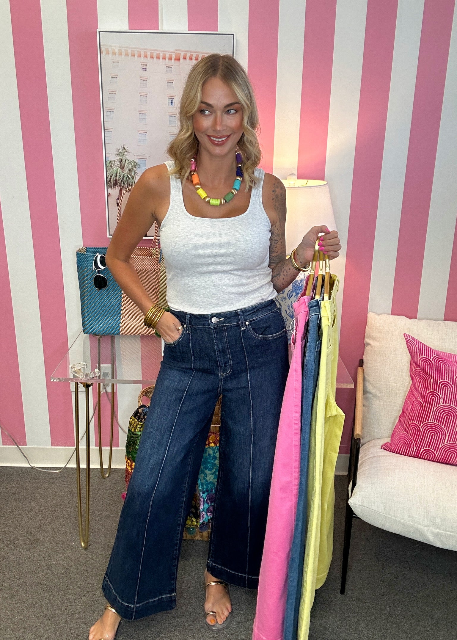 A woman stands in a pink and white striped room holding up a rack of colorful tank tops. She is wearing a white tank top and dark blue wide-leg jeans.