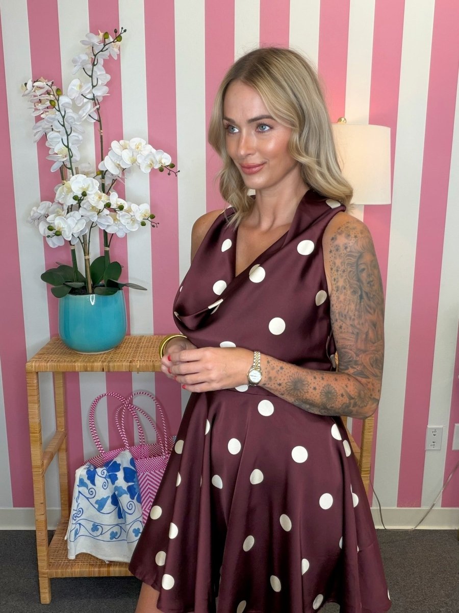 A woman wearing a sleeveless, wrap-style A-line dress in dark burgundy with large white polka dots, standing in front of a pink and white striped wall next to a wooden side table with a turquoise orchid pot.