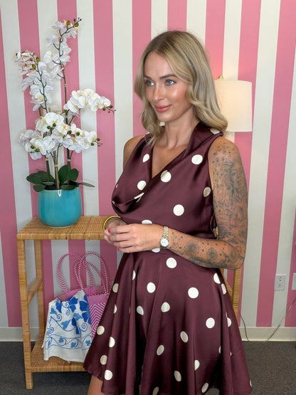 A woman wearing a sleeveless, wrap-style A-line dress in dark burgundy with large white polka dots, standing in front of a pink and white striped wall next to a wooden side table with a turquoise orchid pot.