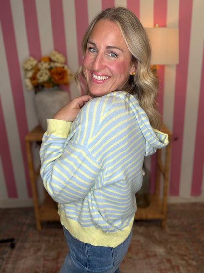 A woman with blonde hair smiling and looking over her shoulder while wearing a light blue and yellow striped hoodie with solid yellow cuffs and hem, set against a pink and white striped wall background.