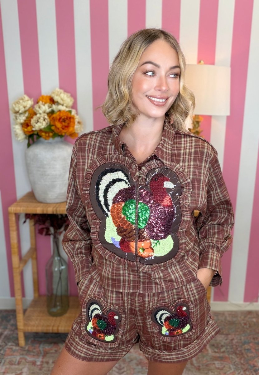 A woman wearing a brown plaid long-sleeve shirt and matching shorts set, both decorated with large sequined turkeys on the front, posed in front of a pink and white striped wall.