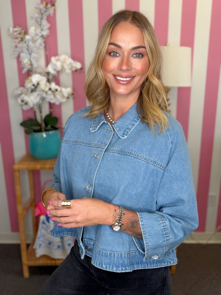 A woman with blonde hair wearing a light blue cropped denim jacket and dark jeans, standing in front of a pink and white striped wall. She is holding a small pink item in her hands, with a wooden side table, white orchids, and a lamp visible in the background.