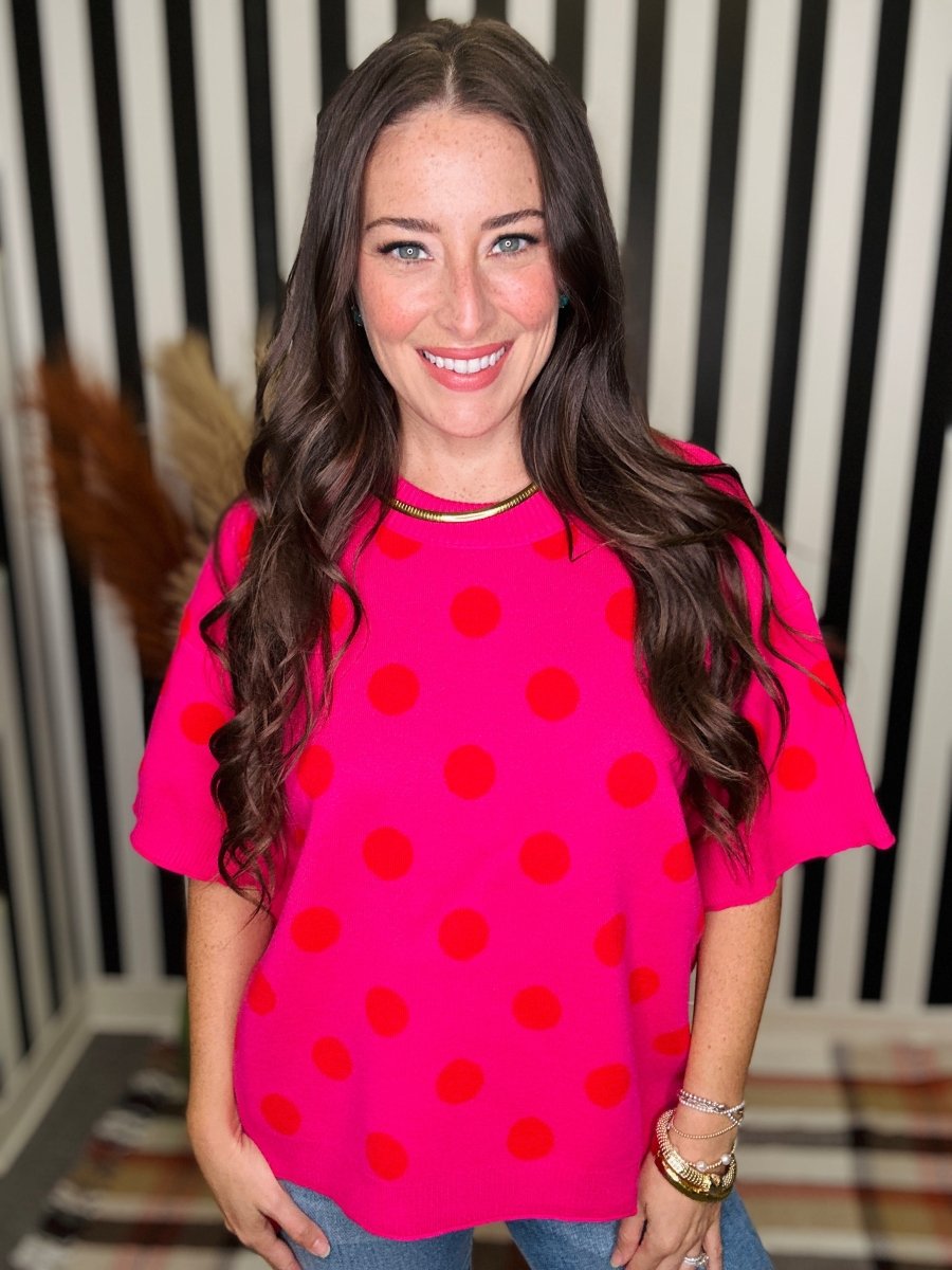A woman with long wavy brown hair smiling, wearing a bright pink short-sleeve top with red polka dots, paired with blue jeans. She is standing in front of a black and white striped background.