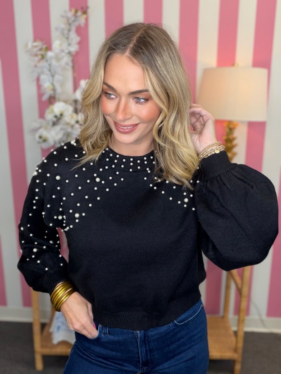 A woman wearing a black knit sweater with white pearl-like embellishments on the chest and shoulders, paired with blue jeans, standing in front of a pink and white striped wall.