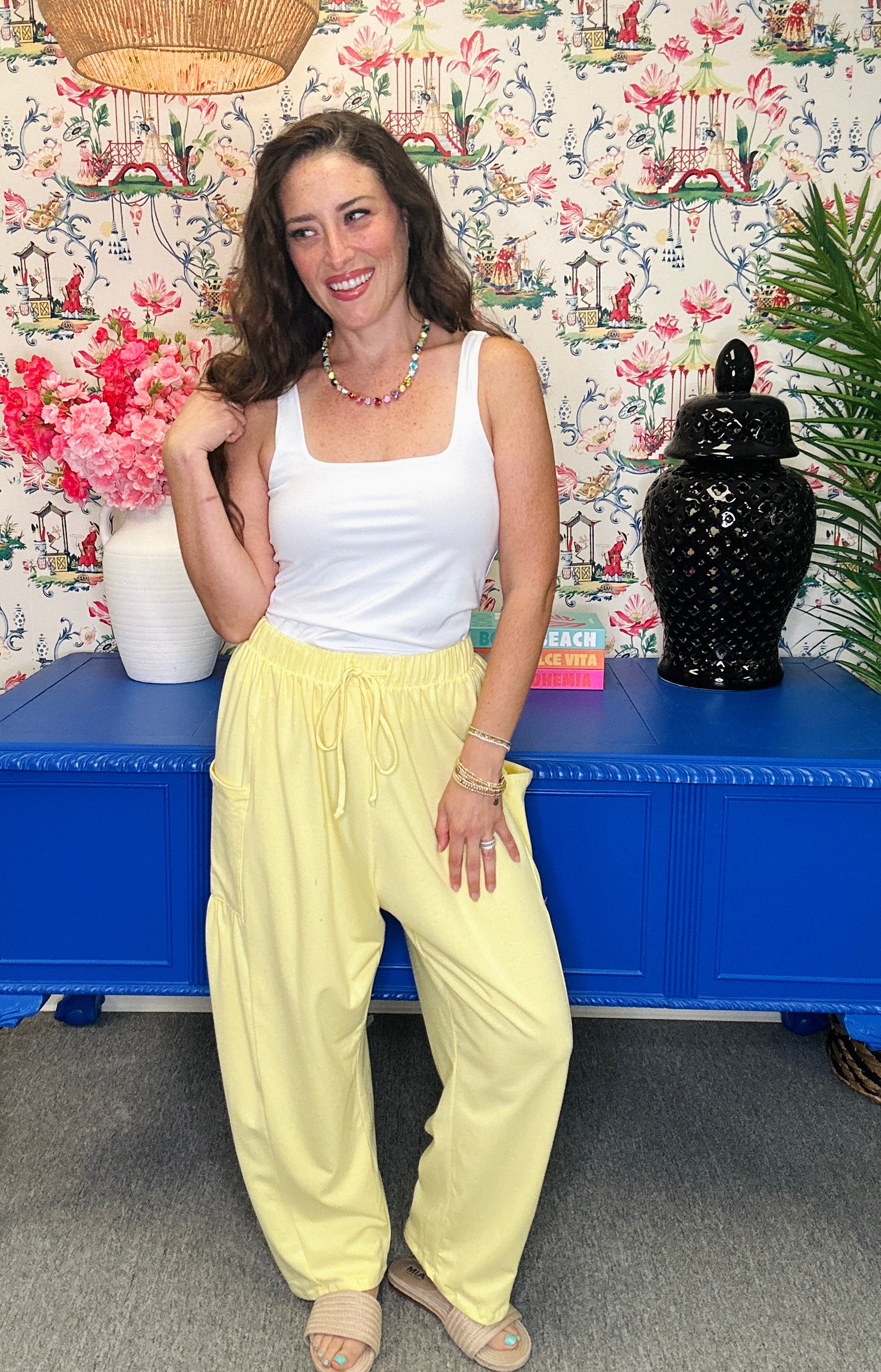 A woman posing in a white tank top and light yellow wide-leg sweatpants, standing in front of a blue cabinet and a floral wallpaper background.