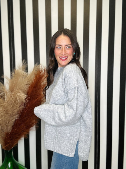 A woman wearing a light gray heathered knit sweater and blue jeans, standing in front of a black and white striped wall with a decorative pampas grass arrangement.