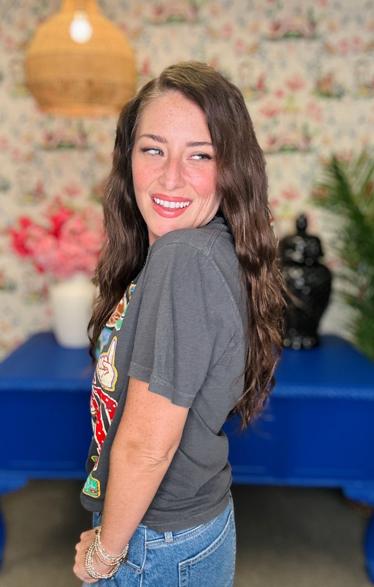 A woman with long brown hair smiling and looking over her shoulder, wearing a dark gray graphic t-shirt and blue jeans in a decorated room with a blue table and floral wallpaper.
