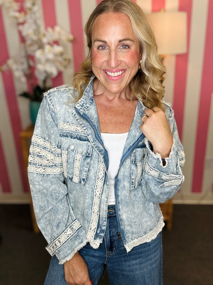 A smiling blonde woman wearing a light-wash acid-wash denim jacket with white lace applique details on the sleeves and front pockets, styled over a white top and blue jeans.