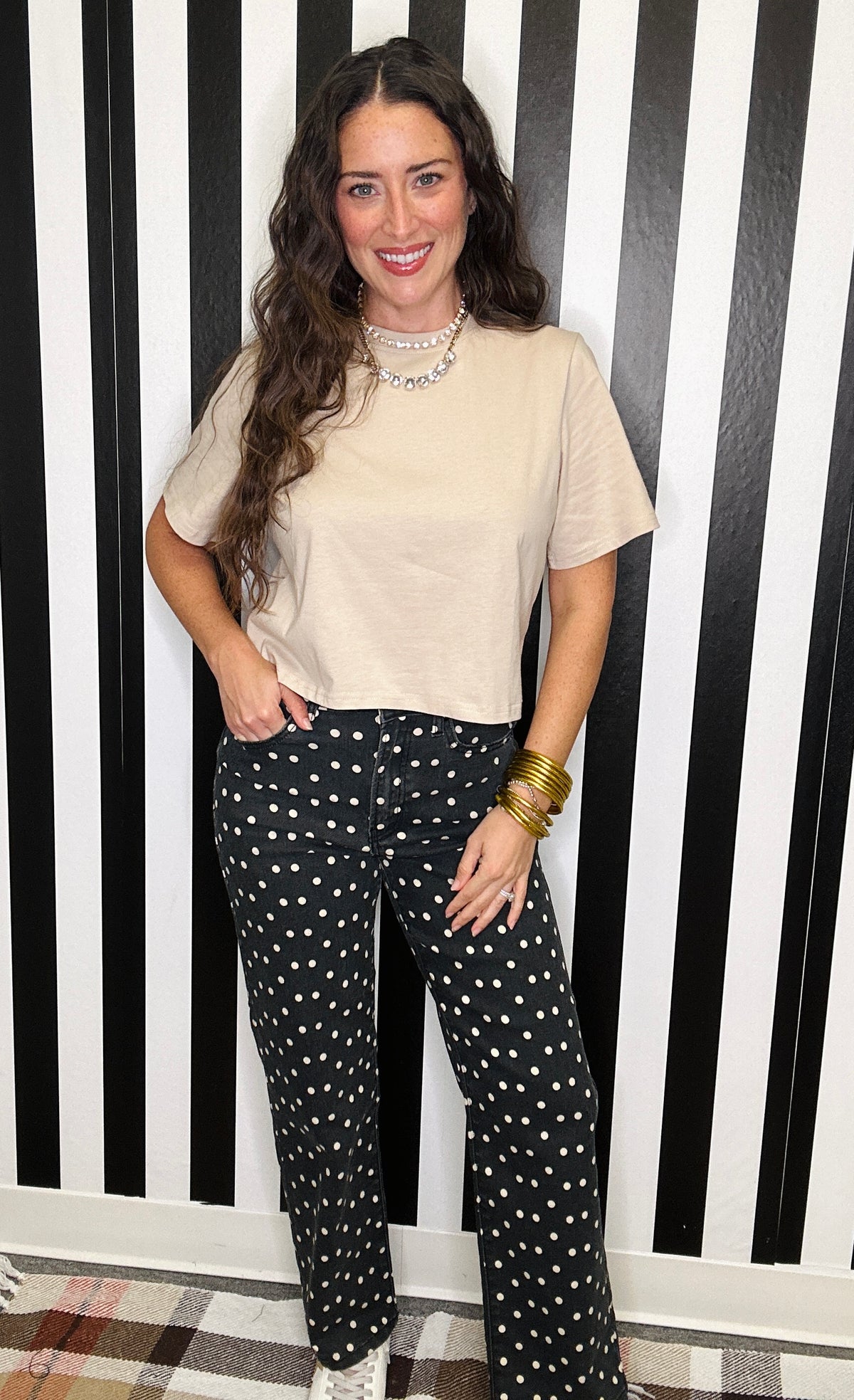 A woman posing in front of a black and white striped wall, wearing a beige short-sleeve crop top, black wide-leg pants with white polka dots, and gold bangles.